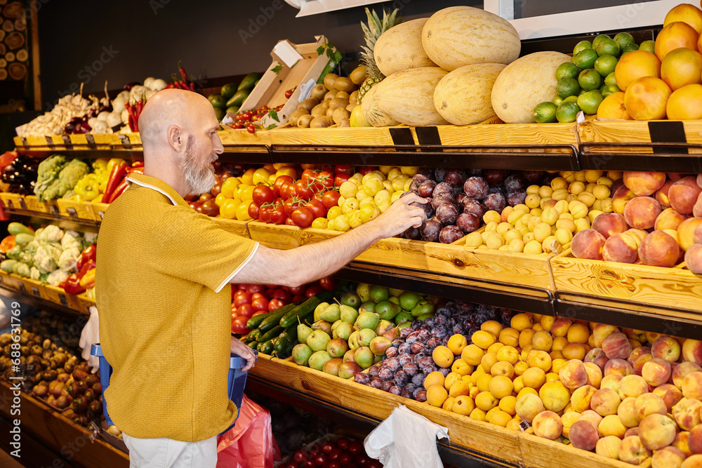 bearded male customer in casual attire with shopping basket choosing fresh plums at grocery store