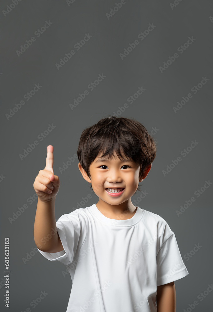 smiling asian boy pointing his finger upward with witty gesture on gray background smiling asian boy pointing his finger upward with witty gesture on gray background