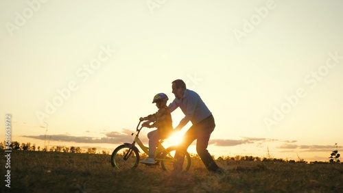 Attentive father silhouette teaches son to ride bike in field at sunset dusk