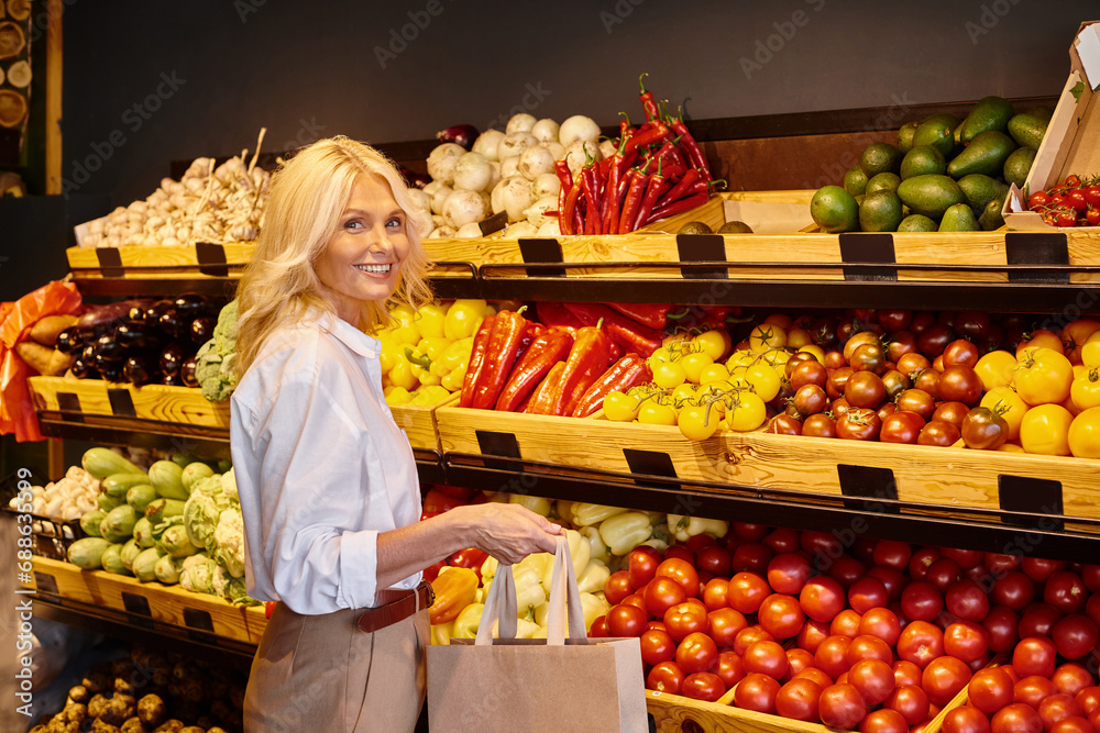 cheerful mature woman in casual attire smiling at camera with vegetable stall on background