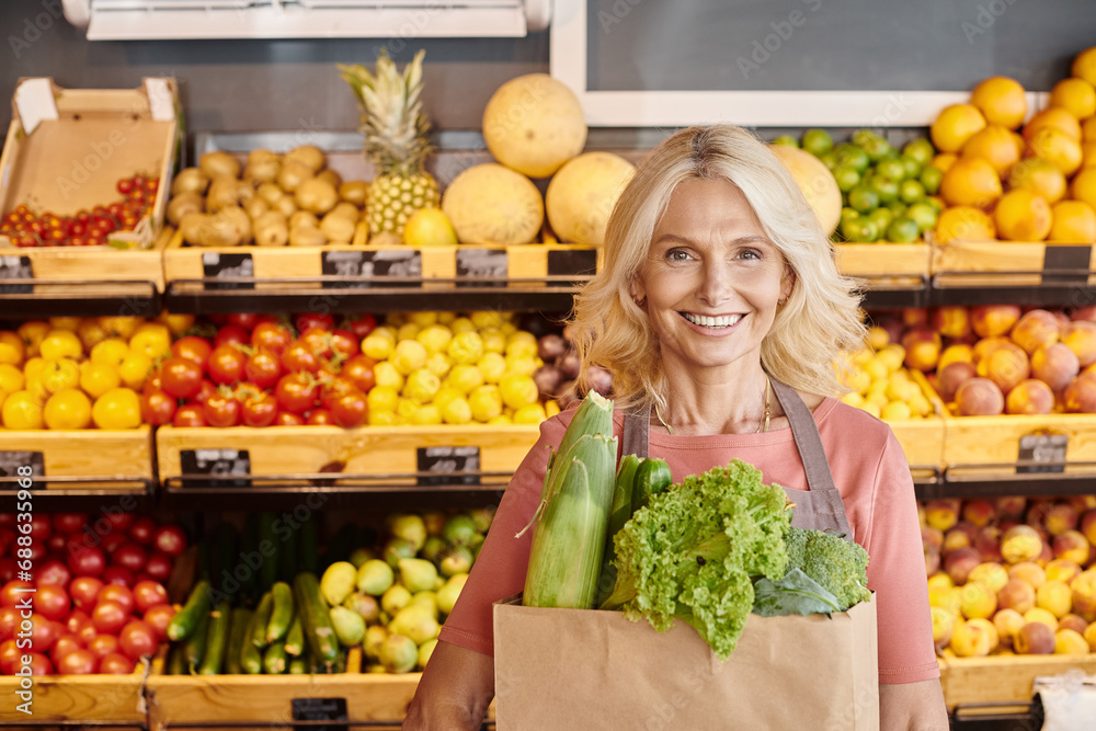 joyous mature seller with paper bag full of greens in hands smiling at camera at grocery store joyous mature seller with paper bag full of greens in hands smiling at camera at grocery store