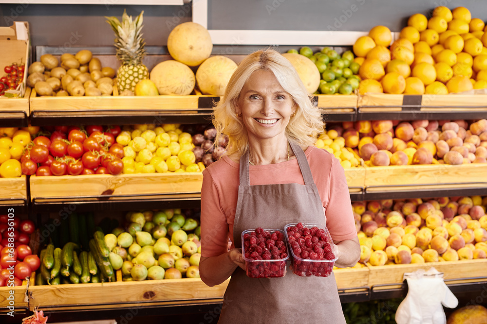 joyous mature saleswoman holding two packs of fresh vibrant raspberries and smiling at camera