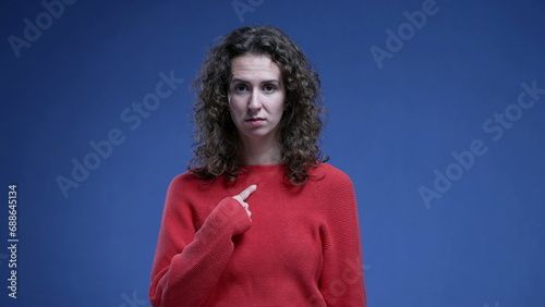 Incredulous woman flabbergasted by accusation, pointing to herself with finger in disbelief standing on blue backdrop. Funny surprised 20s person points with hand to self photo