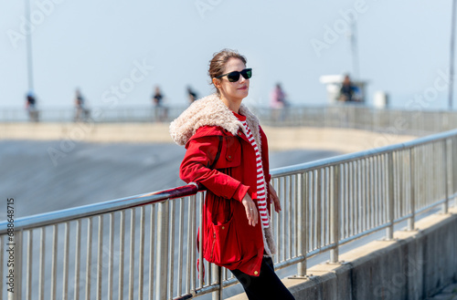 Caucasian woman with red coat and sun glasses lean to fence and look to the right with day light and look cool.