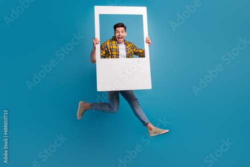 Full body photo of young funky guy jumping with paper photozone window shooting memories at event isolated on blue color background
