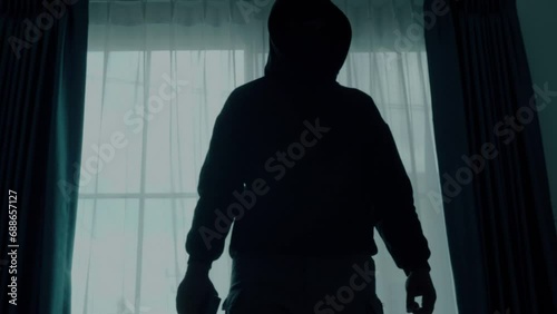 Mysterious man wearing black hoodie holding a pistol wearing a cloth mask covering his face completely in front of the door, Silhouette and dark concept image,threat of firearms,attack or defense.