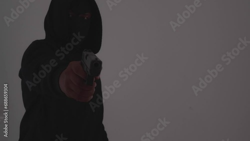 Mysterious man wearing black hoodie holding a pistol wearing a cloth mask covering his face completely in front of the door, Silhouette and dark concept image,threat of firearms,attack or defense.