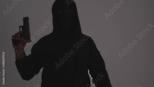 Mysterious man wearing black hoodie holding a pistol wearing a cloth mask covering his face completely in front of the door, Silhouette and dark concept image,threat of firearms,attack or defense.
