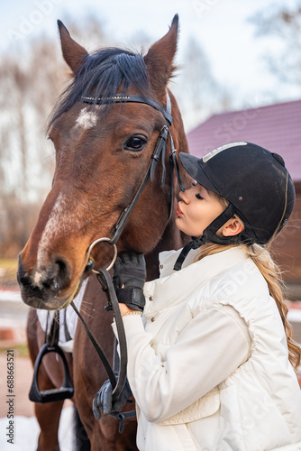 Beautiful blond professional female jockey kissing brown horse on farm. Friendship with horse