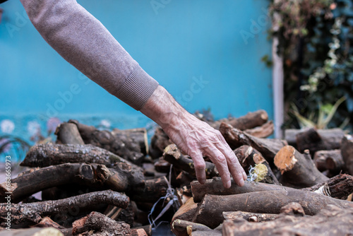 Photo of the hands of an elderly man stacking firewood in front of his house for the cold winter.