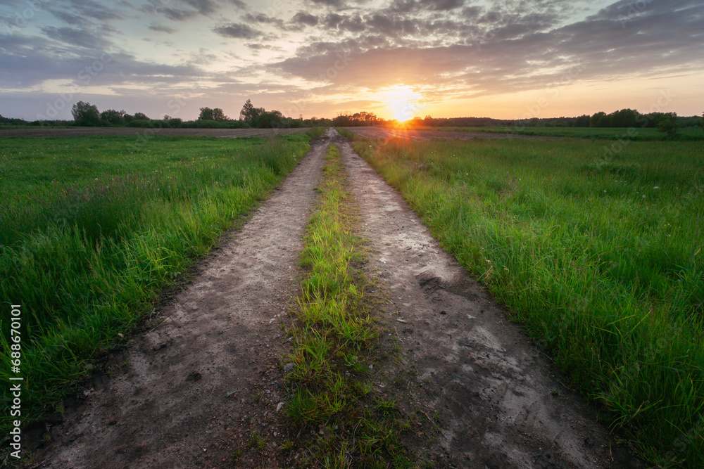 View of a straight and empty dirt road through green meadows with sunset