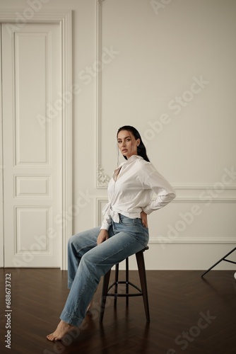 A young brunette woman wearing blue jeans and a white blouse is sitting on a chair in a photo studio. She has a confident and relaxed expression on her face, as she poses for the camera.
