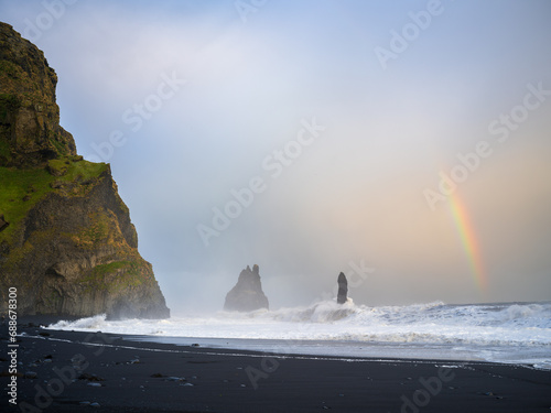 Strong surf at a Reynisfjara black sand beach. Rainbow showing on right side. Reynisdrangar Sea Stacks getting hit by waves. South Coast of Iceland.