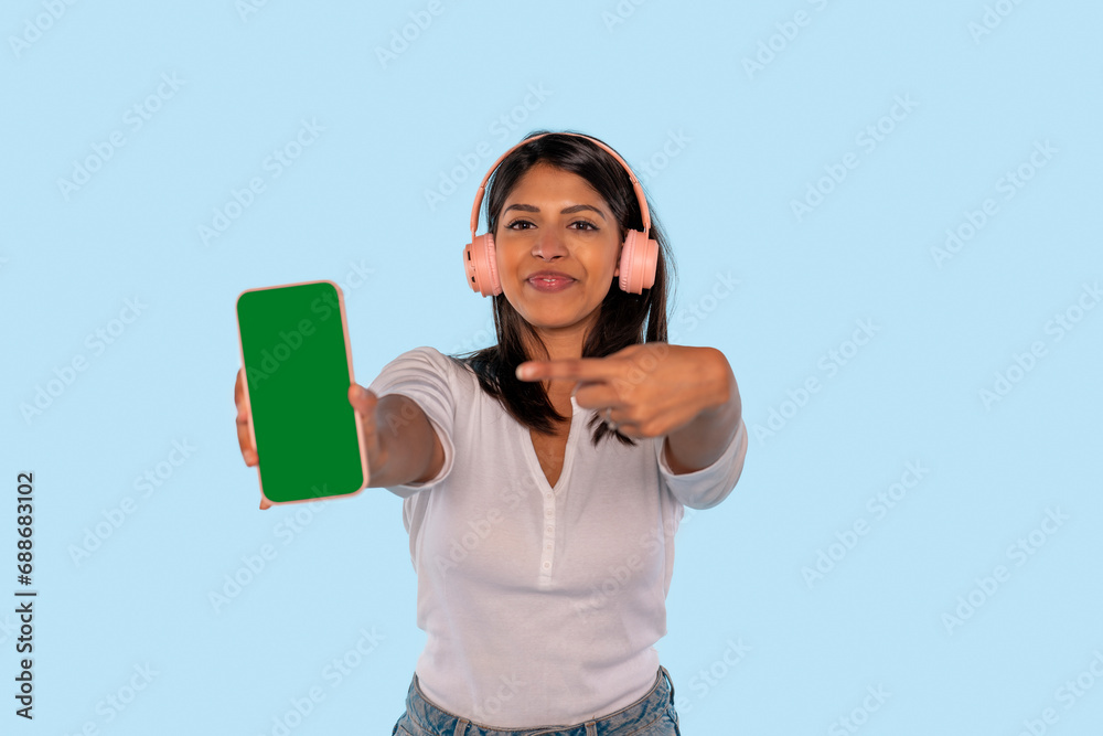 happy indian woman in white shirt and pink headphone showing empty smartphone screen with positive expression, smiles broadly, on blue background. Mobile phone with green screen in female hand. happy indian woman in white shirt and pink headphone showing empty smartphone screen with positive expression, smiles broadly, on blue background. Mobile phone with green screen in female hand.
