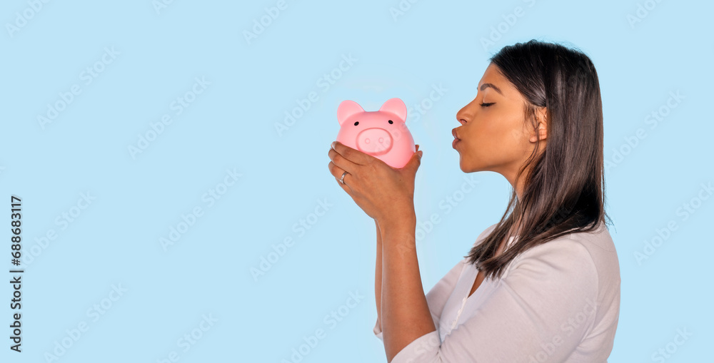 happy indian woman in white shirt kissing piggy bank with savings. Investment, saving money, currency, deposit. Indoor studio shot isolated on blue background. happy indian woman in white shirt kissing piggy bank with savings. Investment, saving money, currency, deposit. Indoor studio shot isolated on blue background.