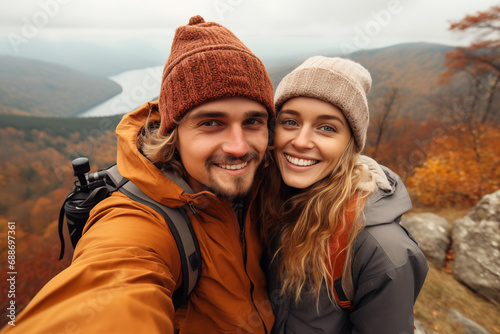 Happy couple taking selfie with mobile phone in autumn forest. Young man and woman in warm clothes and hats standing on top of mountain and looking at camera.