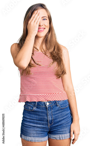 Beautiful caucasian young woman wearing casual clothes covering one eye with hand, confident smile on face and surprise emotion.