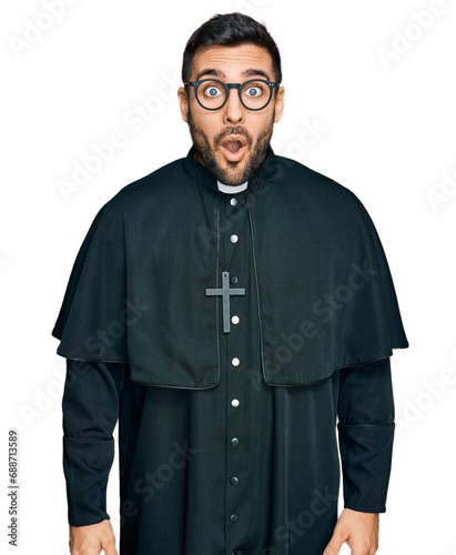 Young hispanic man wearing priest uniform scared and amazed with open mouth for surprise, disbelief face