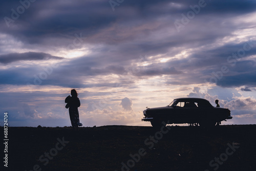 Silhouette of a woman with retro vehicle, standing on the beach. Stormy sea in the background, big waves, Heavy gray rain clouds. Sunset. The concept of travel and recreation on classic cars