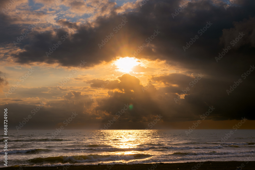Sunset view, seashore, stormy waves, heavy rain clouds on horizon, sun breaking through the cloud like an eye