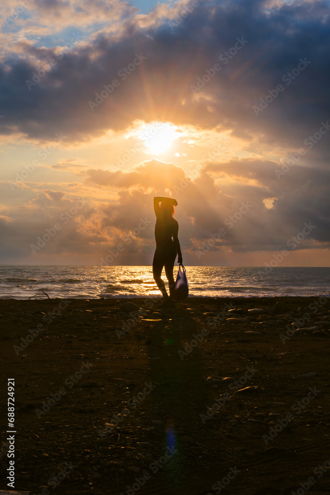Silhouette of a woman with a package at sunset, seashore, in the background stormy waves, heavy rain clouds on horizon, the sun breaks through the cloud like an eye. Vertical photo.