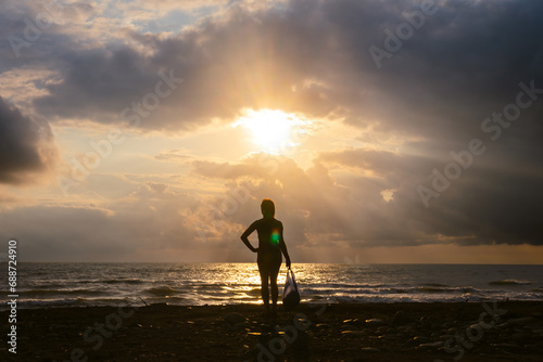 Silhouette of a woman with a package at sunset, seashore, in the background stormy waves, heavy rain clouds on horizon, the sun breaks through the cloud like an eye.