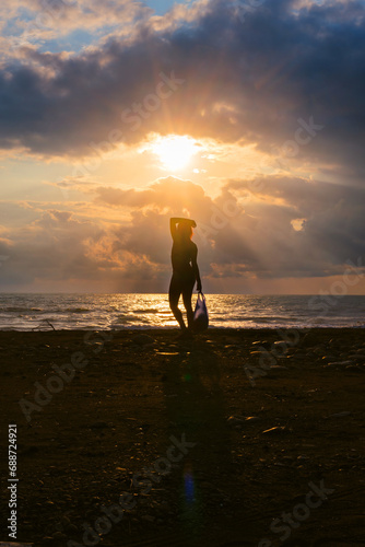 Silhouette of a woman with a package at sunset  seashore  in the background stormy waves  heavy rain clouds on horizon  the sun breaks through the cloud like an eye. Vertical photo.