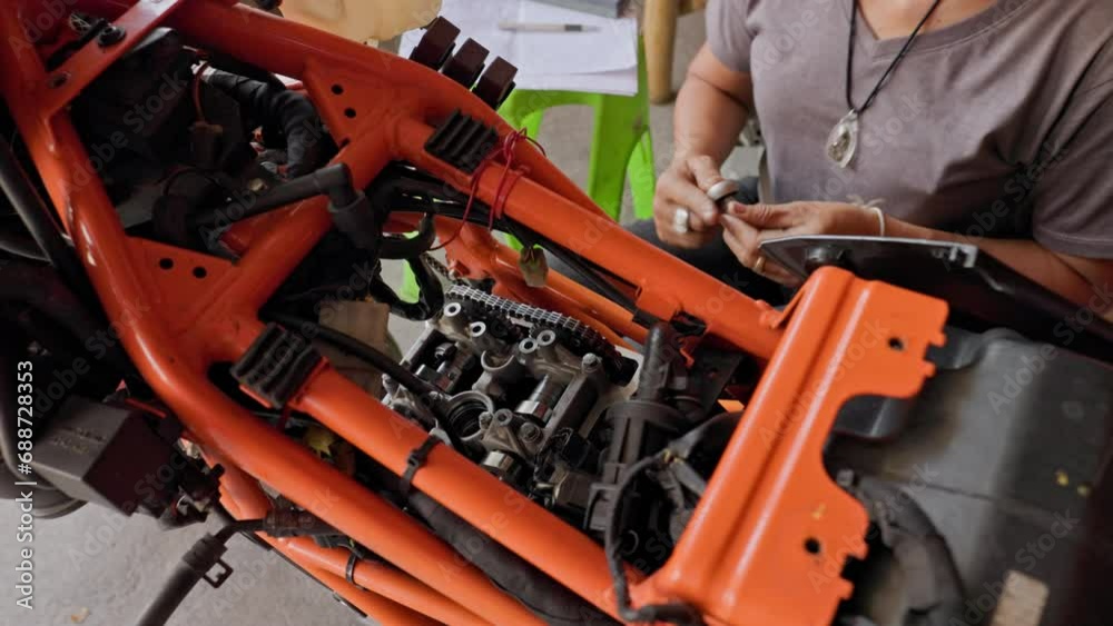 Skillful Restoration: 50 Year Old Asian Woman and the Engine of an Orange Motorcycle Skillful Restoration: 50 Year Old Asian Woman and the Engine of an Orange Motorcycle