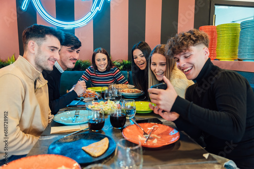 Positive young diverse friends sharing smartphone during dinner in restaurant