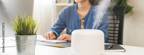 Modern air humidifier device during sitting workplace, happy asian young woman using computer work on internet, enjoying aromatherapy steam scent from essential oil diffuser in room at home office.