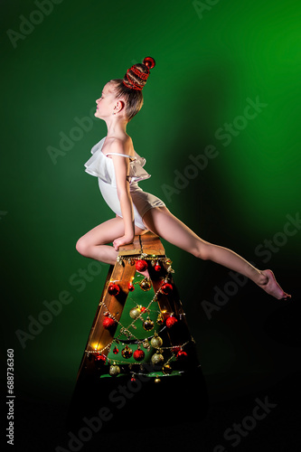 A girl gymnast in a white gymnastic leotard does an acrobatic stretching exercise. Nearby is a decorated wooden Christmas tree. Green background