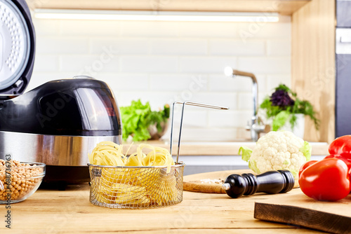 Multi cooker and spaghetti pasta on wooden table in kitchen. Cooking spaghetti or raw nest noodles in a pot. Homemade uncooked ribbon fettuccine. Dry long rolled macaroni close up. photo