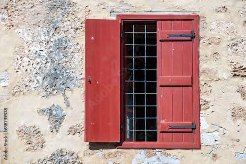 Window with wooden red shutter and metal grid on peeled wall background. Greek architecture.