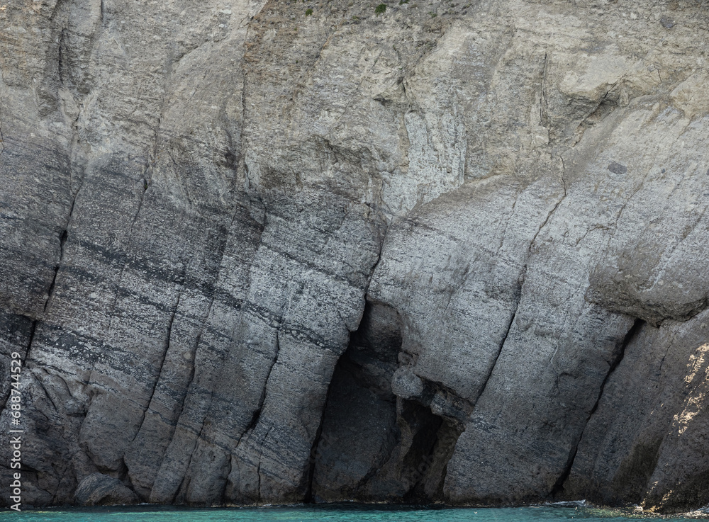 Naklejka premium Weathered seaside rock face texture with blue water. Aged volcanic stone wall surface background pattern with cracks and scratches. Ischia Island, Italy.