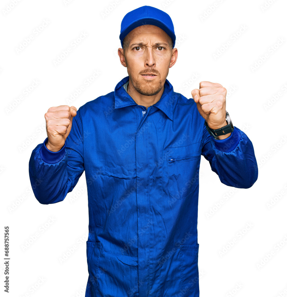 Bald man with beard wearing builder jumpsuit uniform angry and mad raising fists frustrated and furious while shouting with anger. rage and aggressive concept.