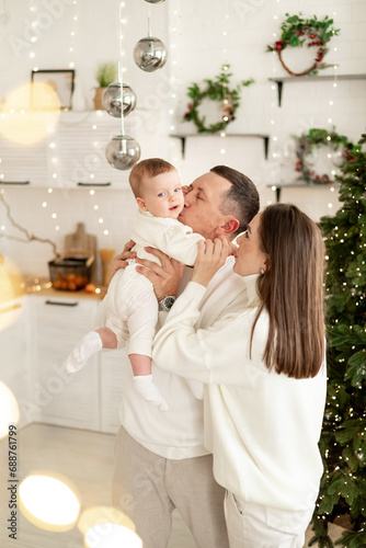 a family with a small child is enjoying the new year at the Christmas tree in the kitchen or in a bright house, smiling, kissing and hugging congratulating each other celebrates the New Year holiday