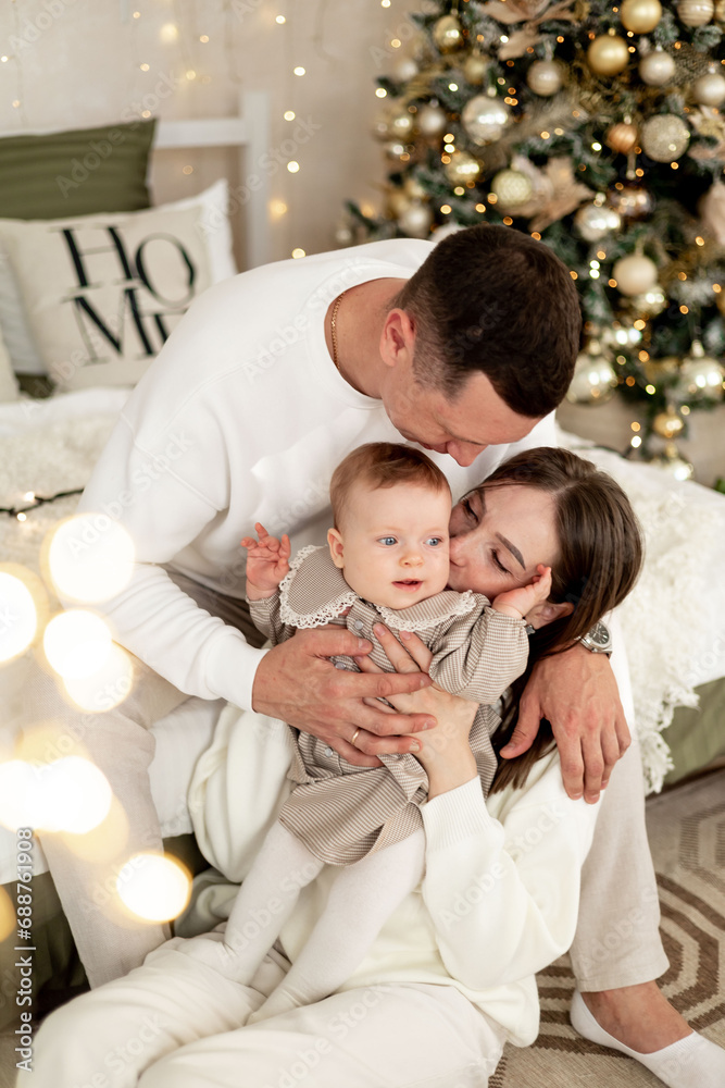 Obraz premium a young family with a small child is enjoying the New year at the Christmas tree on the bed, smiling, kissing and hugging congratulating each other celebrates the holiday