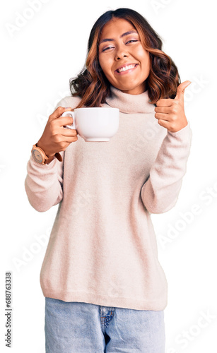 Young beautiful mixed race woman holding a cup of coffee smiling happy and positive, thumb up doing excellent and approval sign