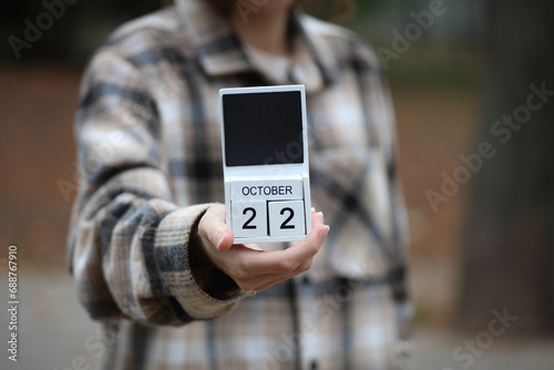 Woman in warm shirt holds calendar with date October 22 outdoors in autumn park