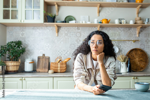 Lonely bored woman sitting alone at home in kitchen, hispanic woman with curly hair depressed thinking holding phone, got bad news message. photo