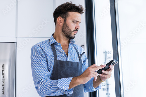 Side view of focused looking man in apron browsing smartphone in daylight near window photo