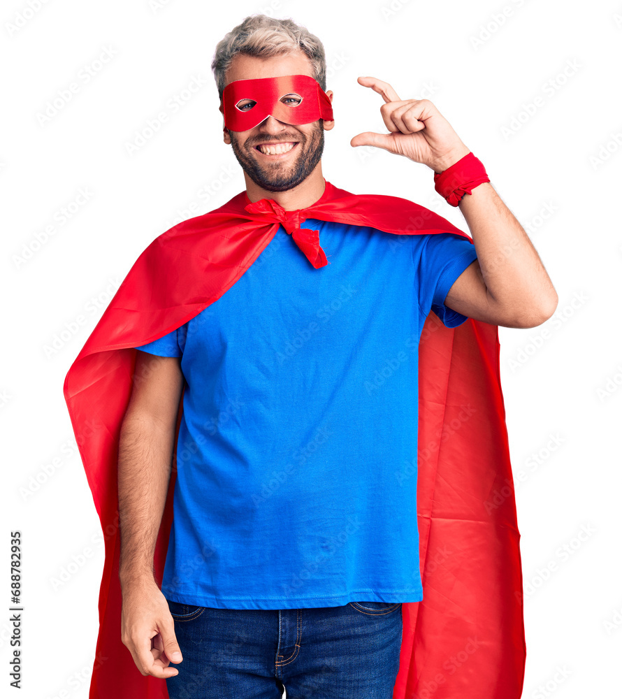 Young blond man wearing super hero custome smiling and confident gesturing with hand doing small size sign with fingers looking and the camera. measure concept. Young blond man wearing super hero custome smiling and confident gesturing with hand doing small size sign with fingers looking and the camera. measure concept.
