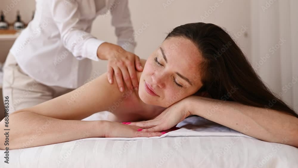 Beautiful topless female customer lying comfortably on bed and receiving back therapy massage from unrecognizable therapist in spa salon Beautiful topless female customer lying comfortably on bed and receiving back therapy massage from unrecognizable therapist in spa salon