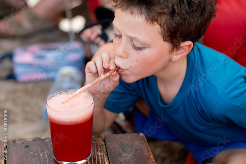 High angle view of little boy in casual clothes drinking fresh watermelon juice in glass with straw while sitting at wooden table in restaurant