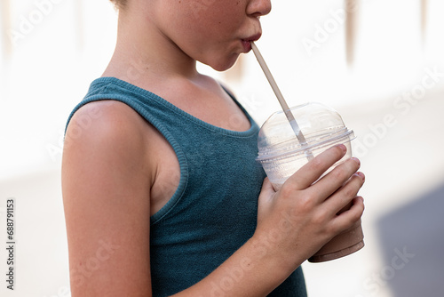 Side view of cropped unrecognizable adorable boy dressed in blue tank top drinking cold coffee with straw while standing on footpath during summer vacation photo