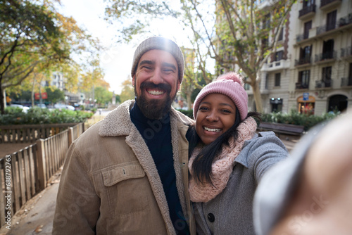 Winter portrait of happy young tourist couple taking selfie outdoors looking at camera in a city. Smiling multi-ethnic people traveling together. Cheerful man and woman posing for photo with phone. photo