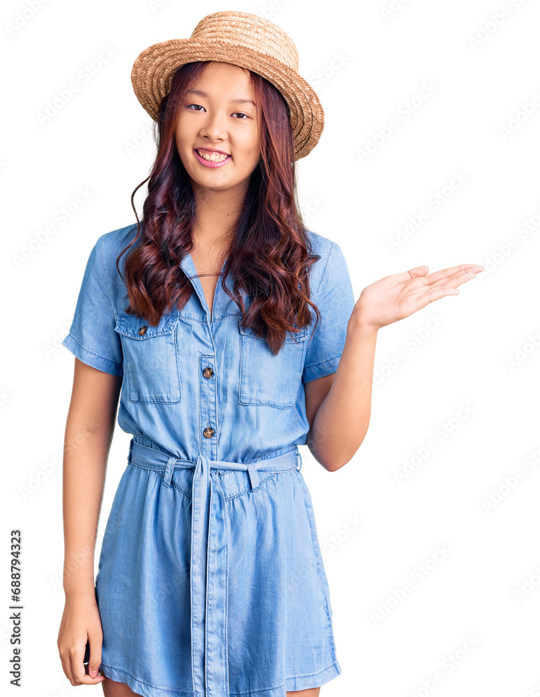 Young beautiful chinese girl wearing summer hat smiling cheerful presenting and pointing with palm of hand looking at the camera. Young beautiful chinese girl wearing summer hat smiling cheerful presenting and pointing with palm of hand looking at the camera.