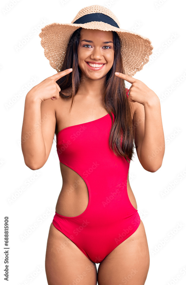 Young beautiful latin girl wearing swimwear and summer hat smiling cheerful showing and pointing with fingers teeth and mouth. dental health concept. Young beautiful latin girl wearing swimwear and summer hat smiling cheerful showing and pointing with fingers teeth and mouth. dental health concept.