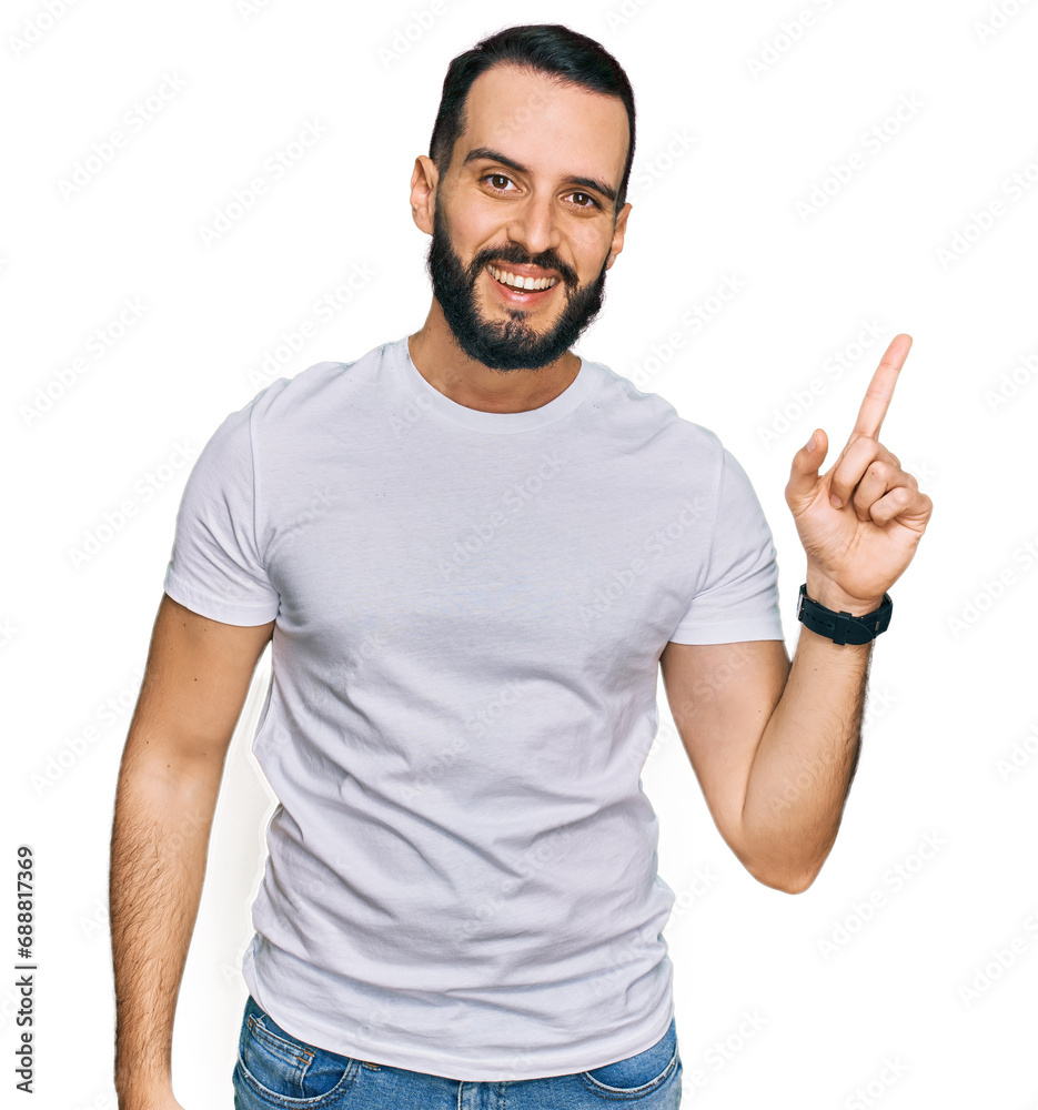 Young man with beard wearing casual white t shirt with a big smile on face, pointing with hand finger to the side looking at the camera. Young man with beard wearing casual white t shirt with a big smile on face, pointing with hand finger to the side looking at the camera.