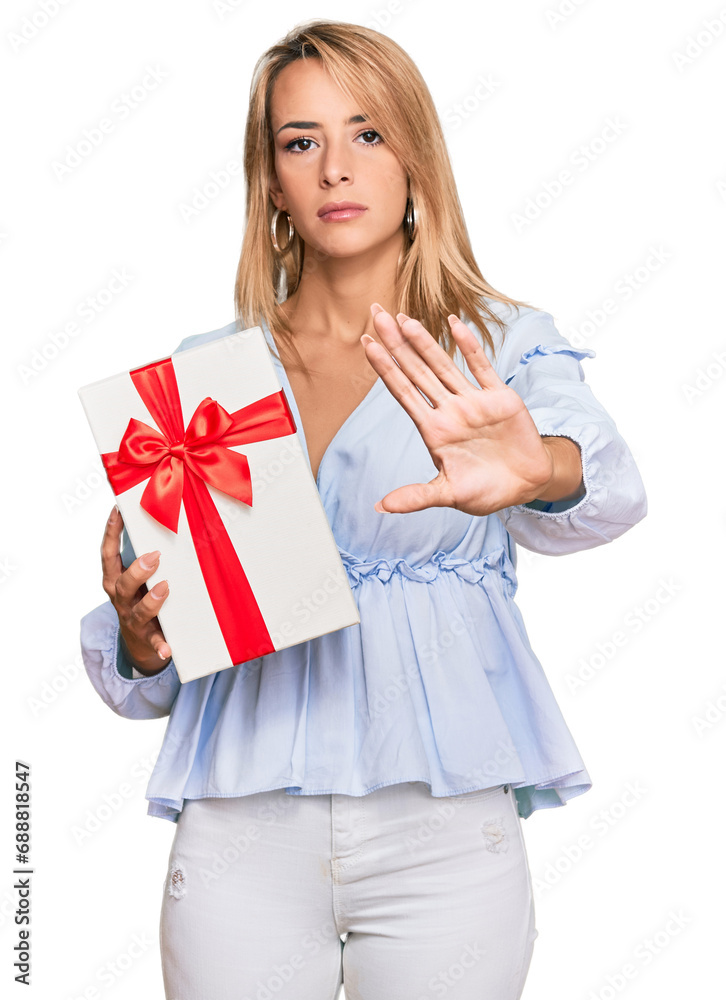 Beautiful blonde woman holding gift with open hand doing stop sign with serious and confident expression, defense gesture Beautiful blonde woman holding gift with open hand doing stop sign with serious and confident expression, defense gesture